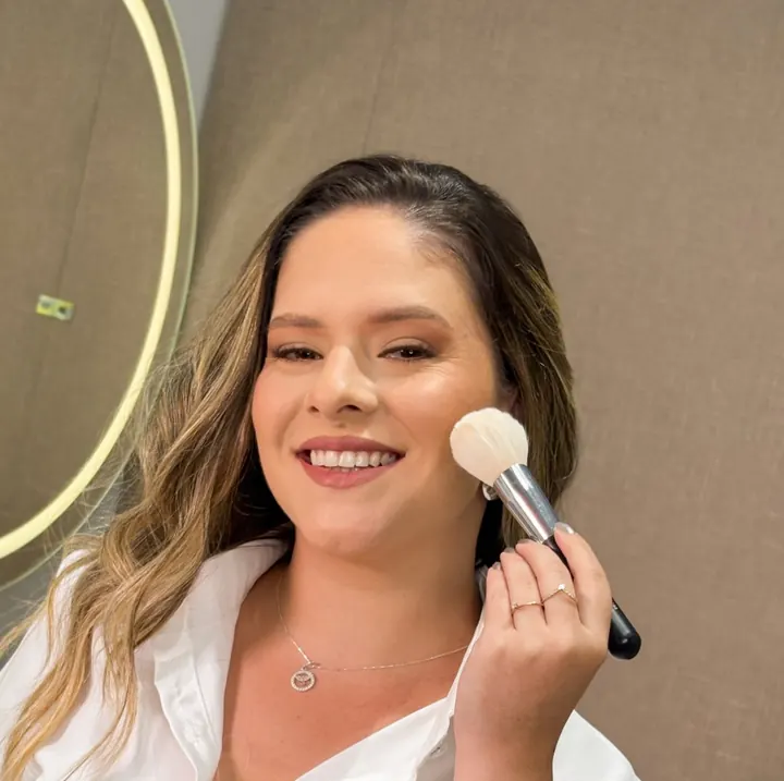 Mulher sorrindo enquanto passa um pincel de pó no rosto, ao lado de um espelho iluminado